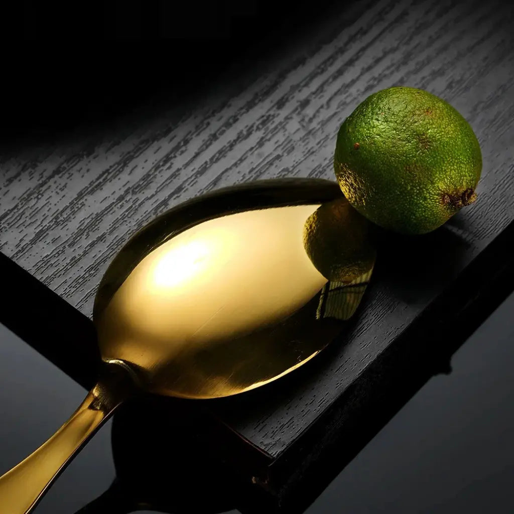 Detalhe refinado da colher dourada do Faqueiro Grand Élysée com fruta cítrica decorativa sobre base escura.